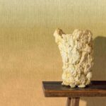 Kunstzinnige vaas op houten tafel voor Elitis Poesies de l’Instant RM 1082 14 behang met warme, gele kleurovergang.