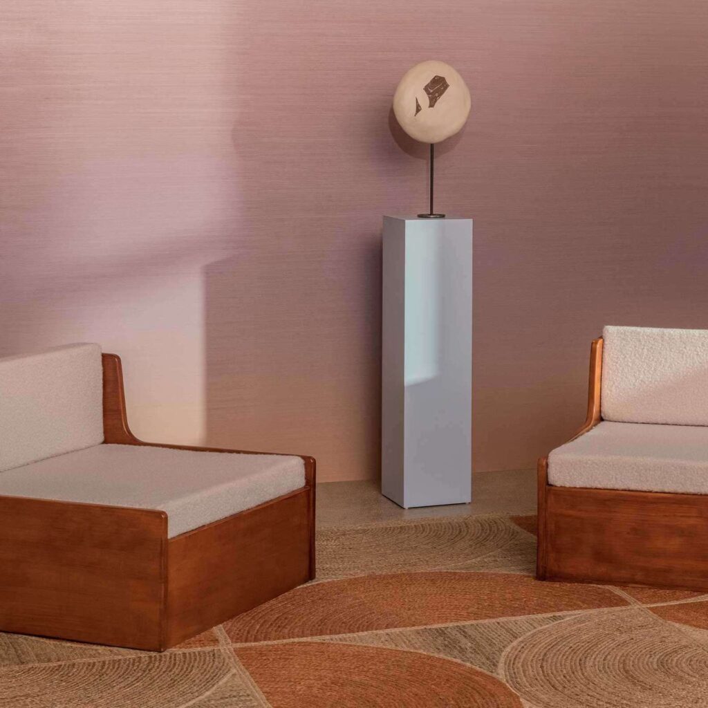 Twee houten loungefauteuils met licht beklede kussens staan tegenover elkaar in een minimalistisch interieur met Elitis Rocky wandbekleding en sculptuur op sokkel.