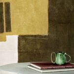 Groene mok en boeken op tafel voor Elitis Sangpi Terres Lointaines behang met abstract patroon.