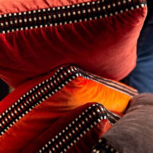 Detailopname van Elitis sierkussens Athena in warme fluweelrode en oranje tinten met een zwart-wit geruit biesje.