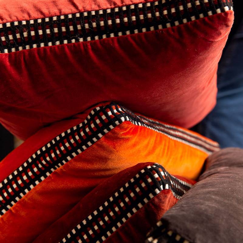 Detailopname van Elitis sierkussens Athena in warme fluweelrode en oranje tinten met een zwart-wit geruit biesje.