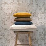Vier Elitis sierkussens uit de Itaque-serie in verschillende kleuren, gestapeld op een houten kruk tegen een decoratieve muur.