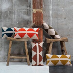 Sierkussens uit de Elitis Minos collectie met geometrische patronen in warme tinten, gestyled op houten krukjes tegen een industriële betonnen muur.