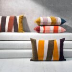 Vier Elitis Nais sierkussens met kleurrijke, diagonale strepen liggen gestyled op en voor een witte bank tegen een betonnen muur.