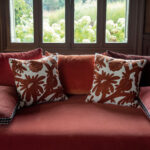 Twee Elitis-sierkussens in rood-wit bloemenmotief liggen op een rood fluwelen bank voor een raam met uitzicht op een tuin.