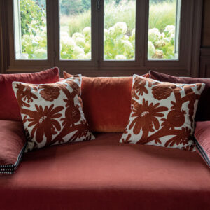 Twee Elitis-sierkussens in rood-wit bloemenmotief liggen op een rood fluwelen bank voor een raam met uitzicht op een tuin.