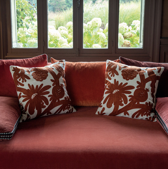 Twee Elitis-sierkussens in rood-wit bloemenmotief liggen op een rood fluwelen bank voor een raam met uitzicht op een tuin.