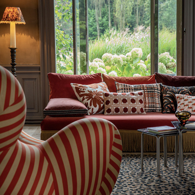 Rode bank met Elitis-sierkussens in diverse patronen in een sfeervolle woonkamer met groot raam en uitzicht op een groene tuin.