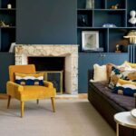 Interieur met een mosterdgele fauteuil en Elitis-sierkussens "Celestine CO 252" in een stijlvolle woonkamer met marmeren haard en donkerblauwe wandkast.
