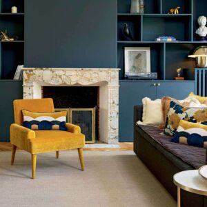 Interieur met een mosterdgele fauteuil en Elitis-sierkussens "Celestine CO 252" in een stijlvolle woonkamer met marmeren haard en donkerblauwe wandkast.