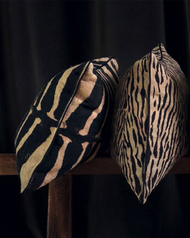 Twee Elitis-sierkussens met zebra- en tijgerprint leunen tegen elkaar op een houten bank voor een donkere achtergrond.