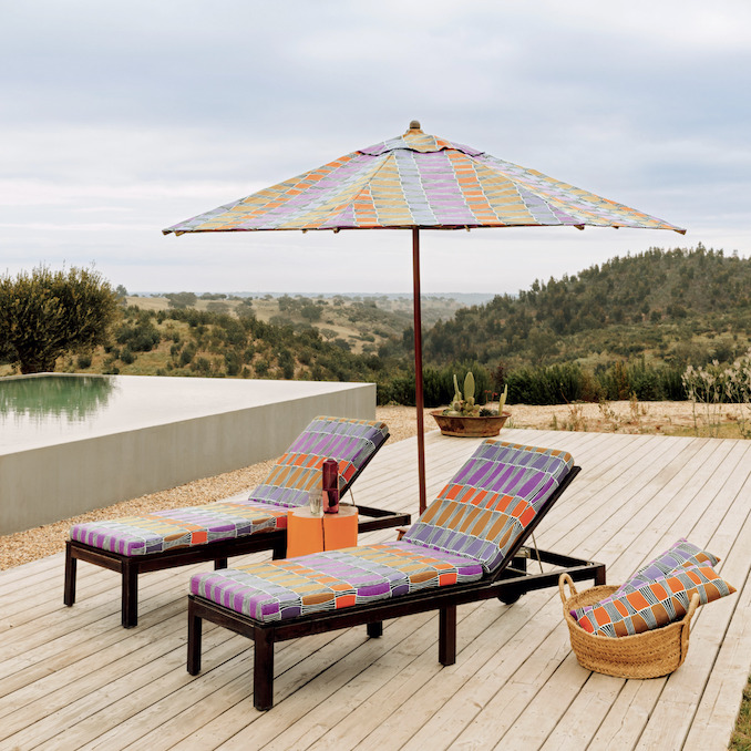 Twee ligstoelen en een parasol bekleed met Elitis-stof Acosta Santa Cruz OD 141 22 staan op een houten terras met uitzicht op een heuvelachtig landschap.