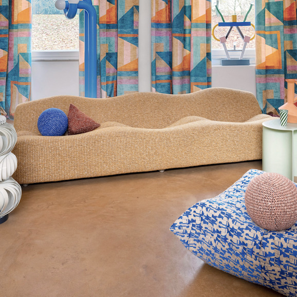 Woonkamer met een golvende beige bank, kleurrijke Elitis Alchimia Giorgio gordijnen en speelse decoratieve kussens.