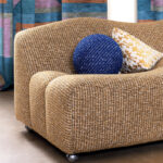 Gevlochten Elitis Alchimia Intreccio-stof op een ronde fauteuil met decoratieve kussens en kleurrijk gordijn op de achtergrond.