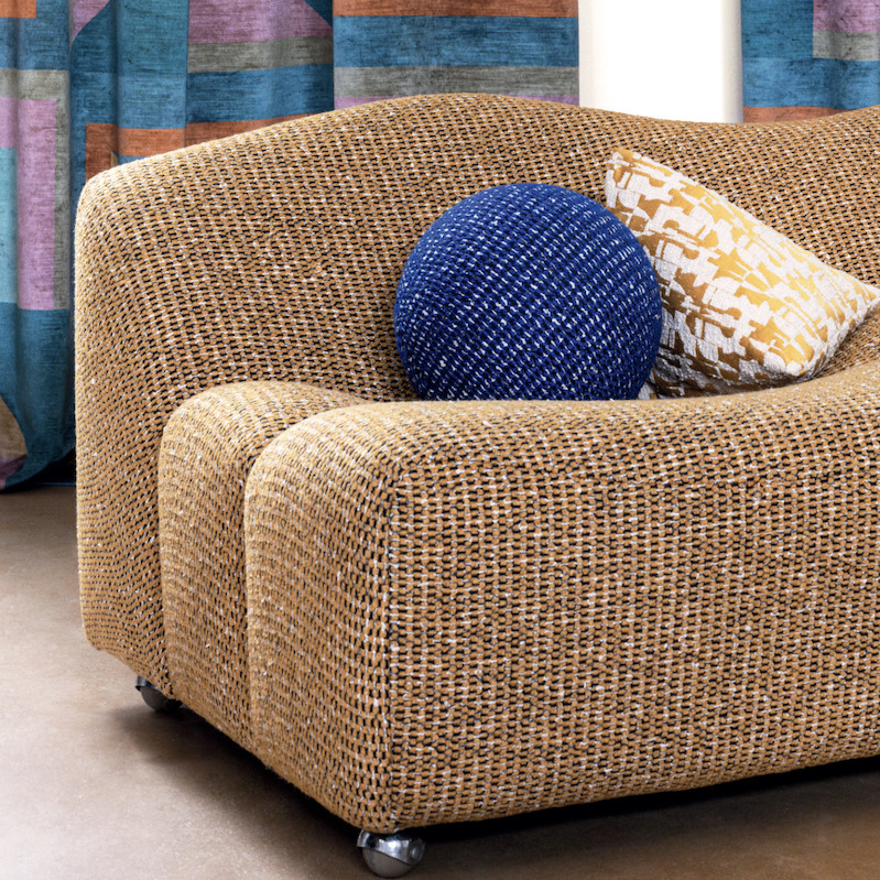 Gevlochten Elitis Alchimia Intreccio-stof op een ronde fauteuil met decoratieve kussens en kleurrijk gordijn op de achtergrond.