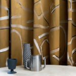 Gouden Elitis-stof Craft Chic Istinto LF 344 21 met abstract patroon, gecombineerd met moderne accessoires op een tafel.