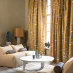 Gezellige zithoek met Elitis gordijnen in het patroon Craft Chic Istinto LF 344 21 in okergeel, gecombineerd met neutrale meubels en moderne accenten.