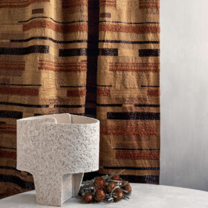 Beige Elitis-stof 'Craft Chic Regina LR 335 24' met geometrisch patroon als gordijn, met een moderne, ruwe keramieken vaas en gedroogde bloemen op een tafel ervoor.