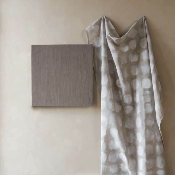 Op een neutrale muur hangt een grijze Elitis-stof Ephemere Full Moon LI 894 05 met witte cirkelpatronen naast een houten paneel.