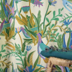Decoratieve Elitis-stof Izamal CN 001 met kleurrijk tropisch patroon van bladeren en vogels, met daarvoor een rotan krukje en een groen kussen.