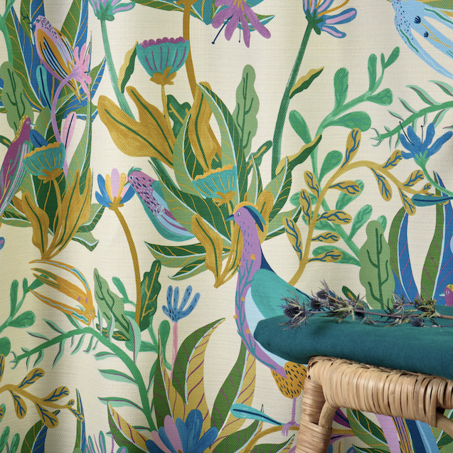 Decoratieve Elitis-stof Izamal CN 001 met kleurrijk tropisch patroon van bladeren en vogels, met daarvoor een rotan krukje en een groen kussen.