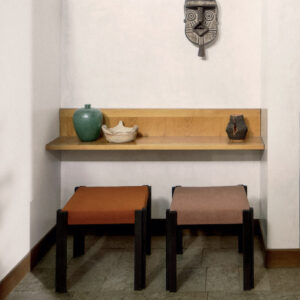 Twee Elitis-stoffen Zelia beklede krukken staan onder een houten wandplank met decoratieve objecten in een minimalistisch interieur.
