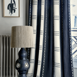Donkerblauwe en crèmekleurige Elitis Escale-gordijnen met etnisch patroon naast een decoratieve lamp en abstracte wandkunst in een stijlvolle interieurhoek.