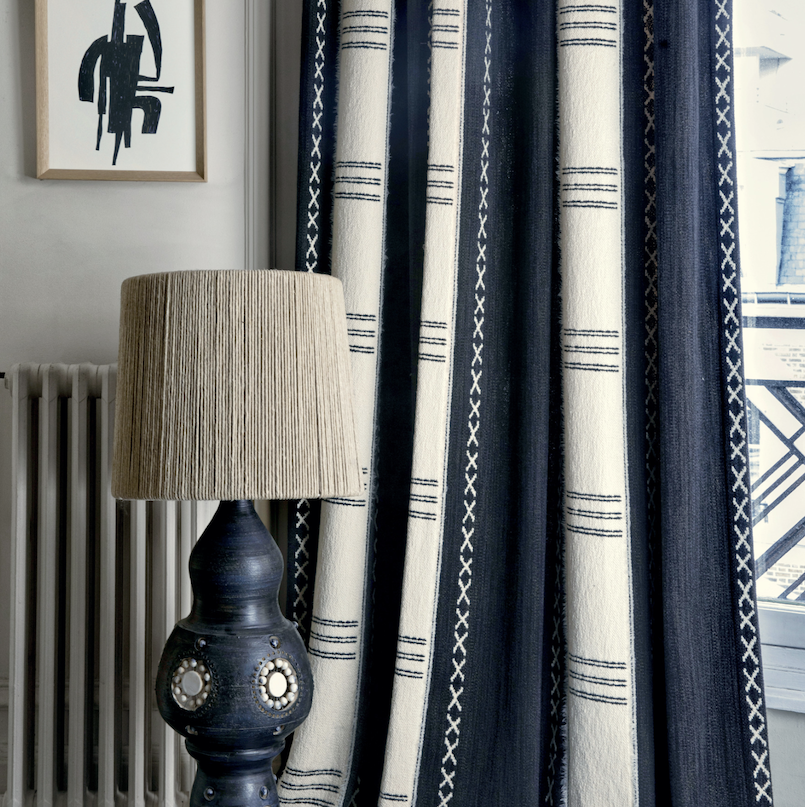 Donkerblauwe en crèmekleurige Elitis Escale-gordijnen met etnisch patroon naast een decoratieve lamp en abstracte wandkunst in een stijlvolle interieurhoek.
