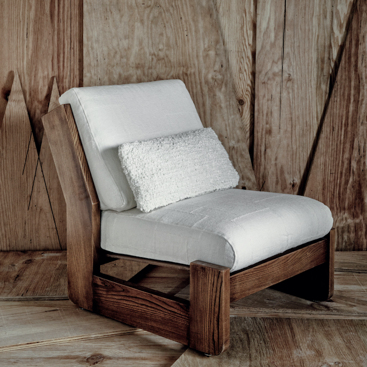 Lage houten loungefauteuil met wit Elitis Esprit LI 871 01 stoffering en rechthoekig sierkussen, geplaatst tegen een houten wand.