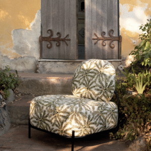 Loungestoel met Elitis outdoor-stof met bladprint staat in een mediterrane tuin voor een verweerde gele gevel met houten luiken.