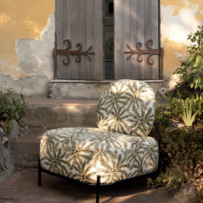 Loungestoel met Elitis outdoor-stof met bladprint staat in een mediterrane tuin voor een verweerde gele gevel met houten luiken.