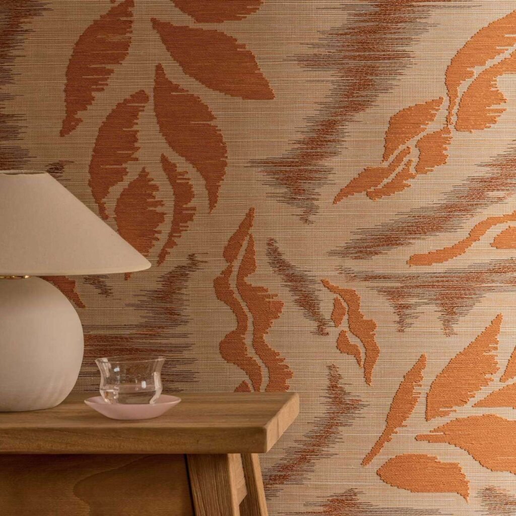 Beige muur met Elitis Tribus Iskandar RM 1097 35 behang met oranje bladmotief, naast een houten tafel met een lamp en een glazen schaaltje.