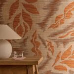 Beige muur met Elitis Tribus Iskandar RM 1097 35 behang met oranje bladmotief, naast een houten tafel met een lamp en een glazen schaaltje.