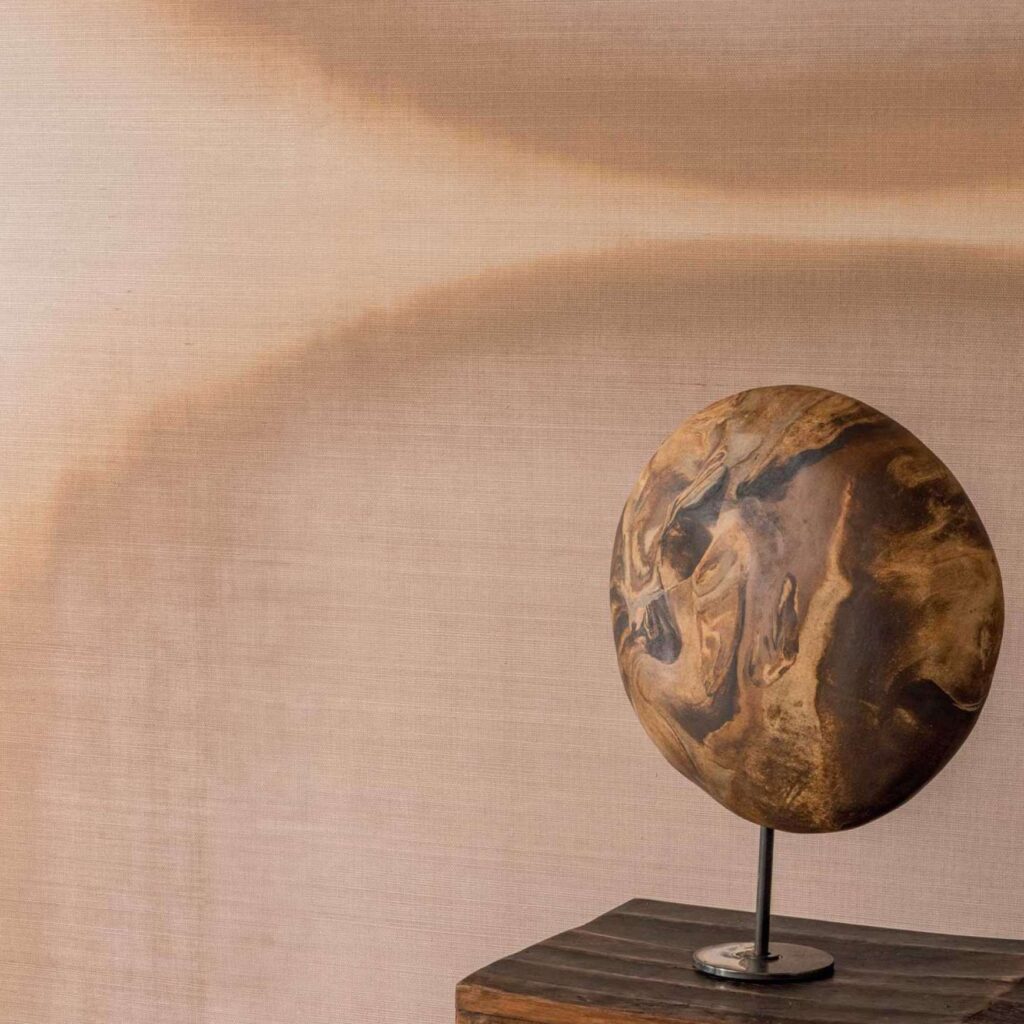 Detail van Elitis Tribus Itza RM 1081-04 behang met aardetinten en een decoratief rond object op een houten tafel.