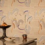 Decoratieve sfeerfoto met Elitis Tribus Lazgi RM 1100 01 behang met abstracte vormen en kleuraccenten op een beige achtergrond, met een schaal fruit en keramiek op een tafel op de voorgrond.