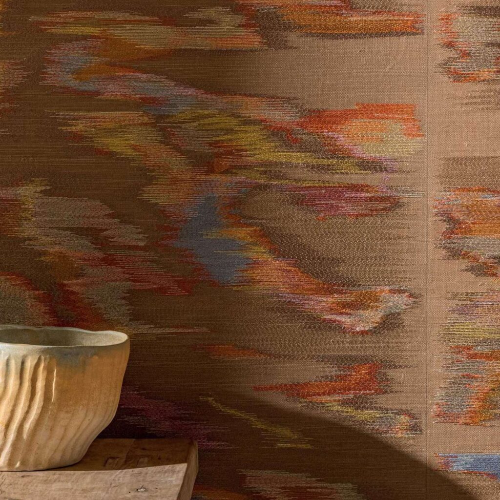 Wandbekleding Elitis Tribus Samarkand met een abstract, kleurrijk patroon in warme aardetinten en een handgemaakte keramieken schaal op een houten oppervlak.
