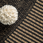 Detailopname van het Elitis vloerkleed Havane met een grafisch patroon in zwart en beige en een decoratieve witte bloem.