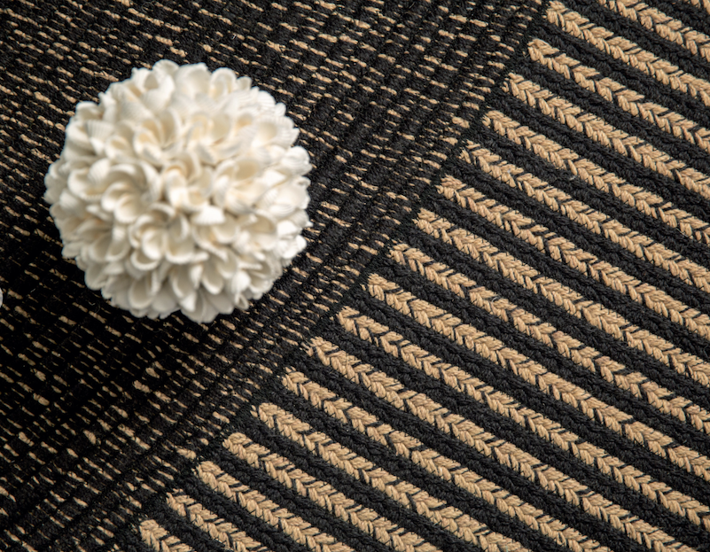 Detailopname van het Elitis vloerkleed Havane met een grafisch patroon in zwart en beige en een decoratieve witte bloem.