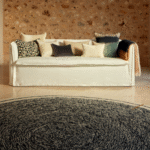 Woonkamer met Elitis vloerkleed Oscar TA 131 02 07 in zwart, voor een beige bank met sierkussens tegen een stenen muur.