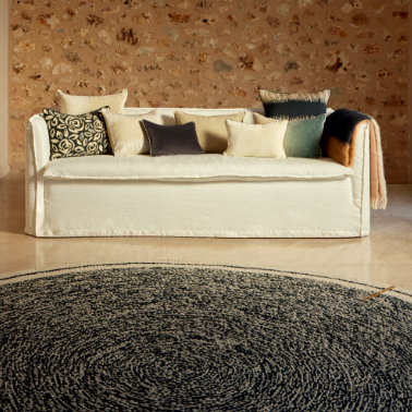 Woonkamer met Elitis vloerkleed Oscar TA 131 02 07 in zwart, voor een beige bank met sierkussens tegen een stenen muur.