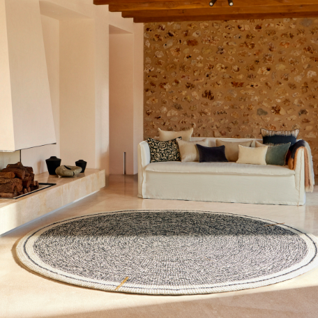 Rond Elitis vloerkleed Oscar TA 131 02 07 in een stijlvolle woonkamer met stenen muur en lichte sofa.