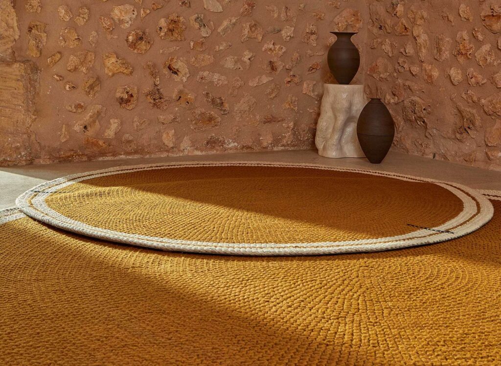 Rond okergeel Elitis vloerkleed Oscar TA 131 02 07 met witte rand in een rustiek interieur met stenen muur en decoratieve vazen.