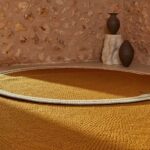 Rond okergeel Elitis vloerkleed Oscar TA 131 02 07 met witte rand in een rustiek interieur met stenen muur en decoratieve vazen.