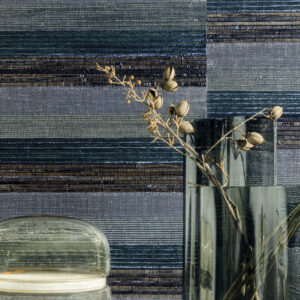 Glasvazen met gedroogde takken staan voor een wand bekleed met Elitis SOLO silver wandbekleding in horizontale banen van blauw, zilver en groen.