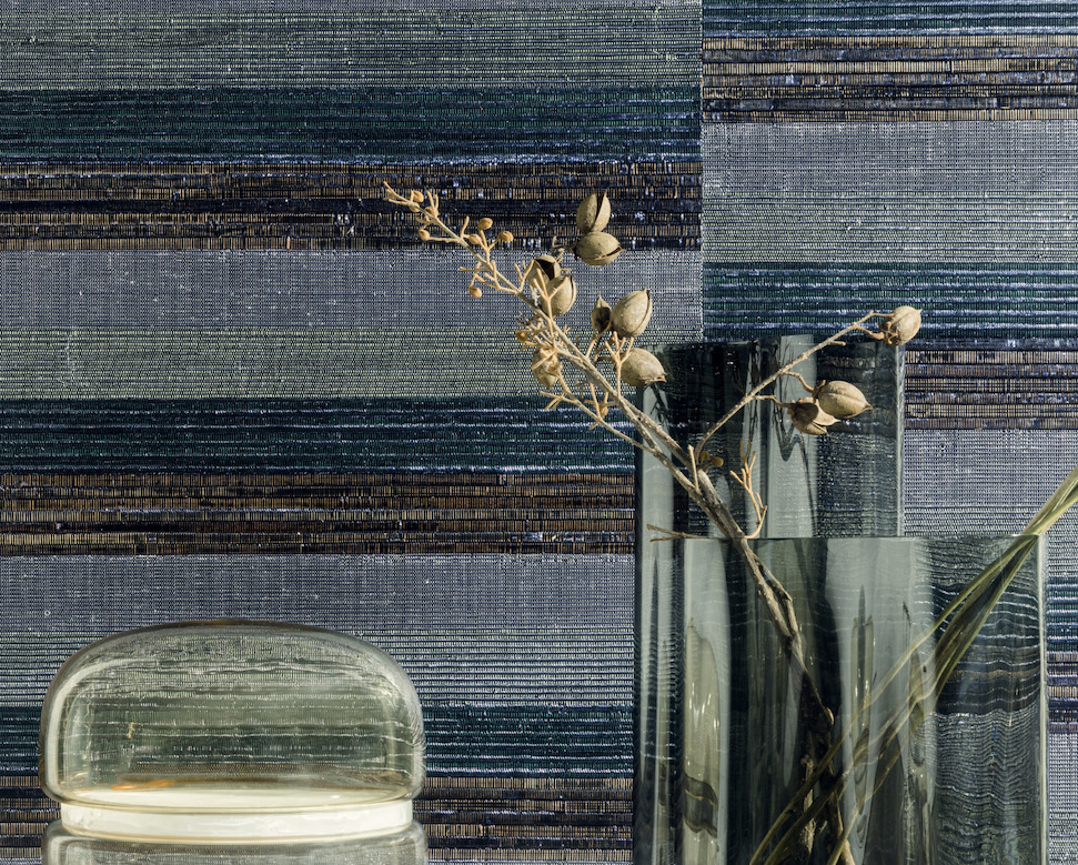 Glasvazen met gedroogde takken staan voor een wand bekleed met Elitis SOLO silver wandbekleding in horizontale banen van blauw, zilver en groen.