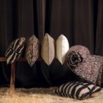 Zes Eltis-sierkussens en een opgerolde deken met tijger- en dierenprints liggen gestyled op een houten bank voor een donkere gordijnachtergrond.
