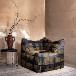 Moderne fauteuil met Eltis-stof in blauw en okergeel ruitpatroon in een sfeervolle, artistiek ingerichte hoek met ruwe muren en een decoratieve vaas op een bijzettafel.