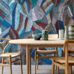 Houten eettafel met keramische decoratie en gevlochten stoelen, tegen een kleurrijk grafisch behang uit de Essentials Les Tricots 73100 collectie.