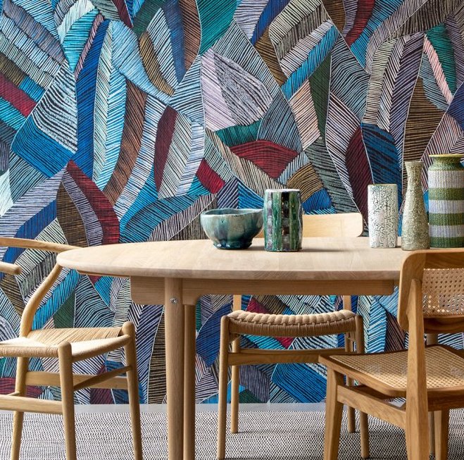 Houten eettafel met keramische decoratie en gevlochten stoelen, tegen een kleurrijk grafisch behang uit de Essentials Les Tricots 73100 collectie.