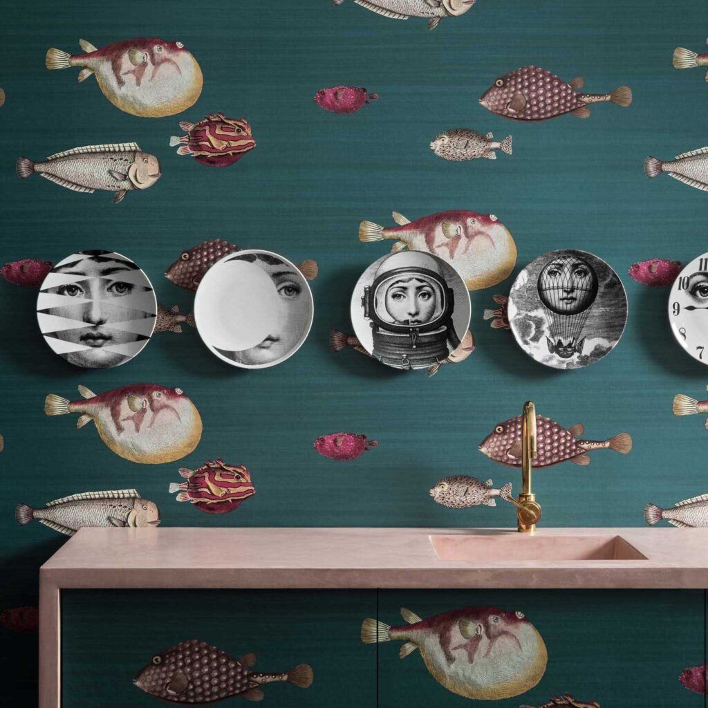 Wand met Fornasetti Acquario-behang met kleurrijke vismotieven en decoratieve borden met surrealistische gezichten boven een roze wastafel.